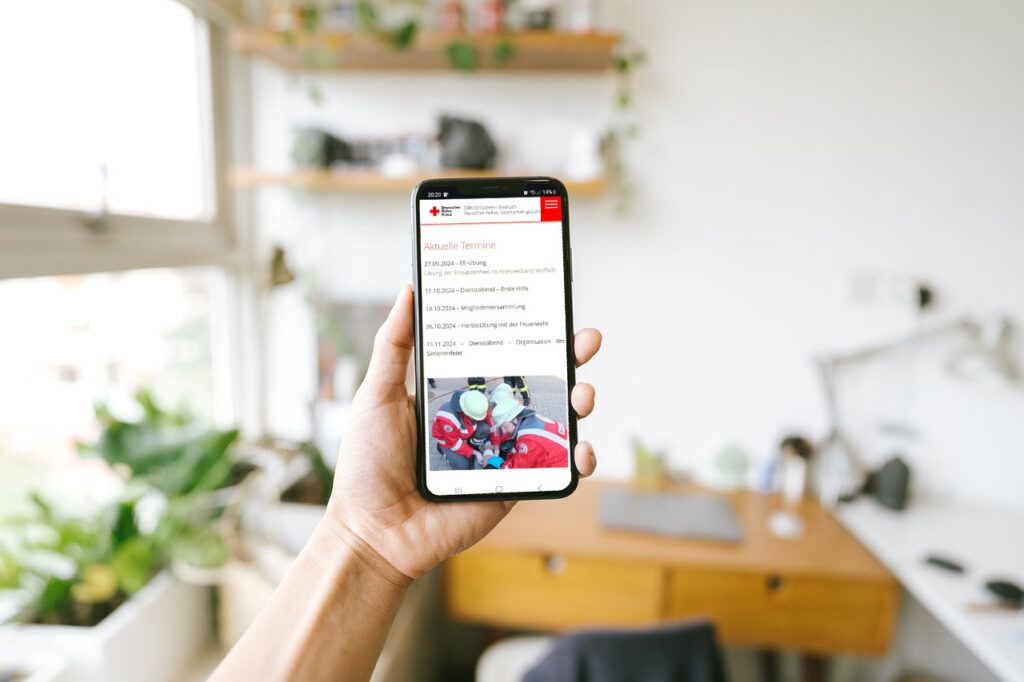 Das Bild zeigt die Hand einer Frau, die ein Handy in der Hand hält. Darauf ist ein Ausschnitt der Website des DRK OV Steinach zu sehen.